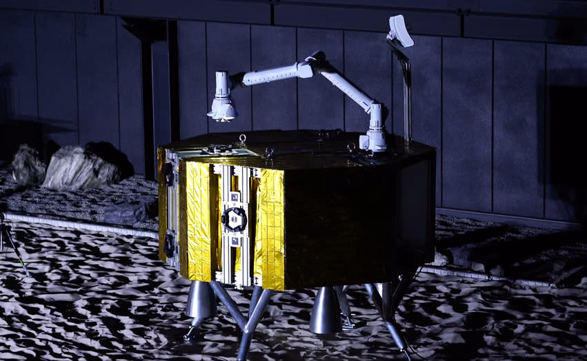  A gold-foil-wrapped octagonal lunar probe is landed on a grey surface. A white robotic arm is attached to the probe's flat top, next to an antenna. 