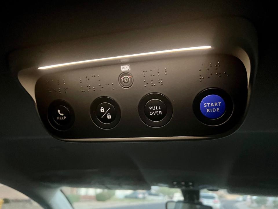 The buttons on the ceiling of the car, including black buttons for pull over, call assistance, and lock/unlock. A blue button starts the ride.