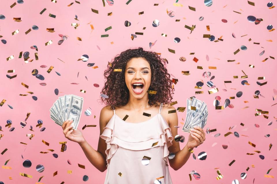A happy person holds up handfuls of cash while being showered by confetti.