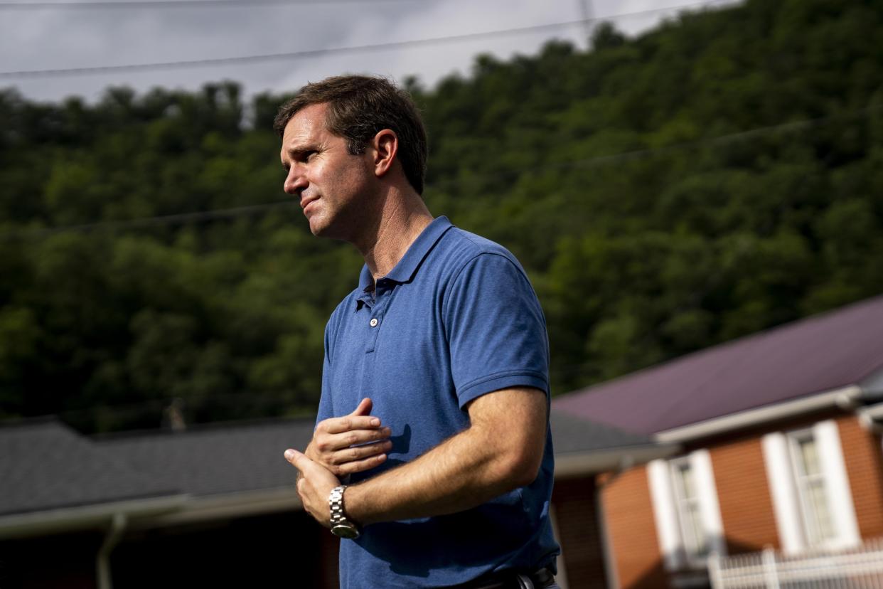 Beshear in a polo shirt with his arms in front of his torso, with leafy green trees and a one-story brick house behind him.