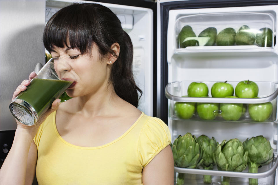 Person drinking a green smoothie with a fridge filled with green apples in the background