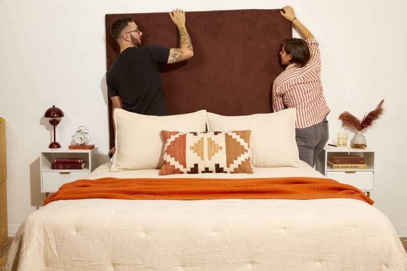 Head on shot of two people hanging the brown DIY headboard onto the wall behind the made bed.