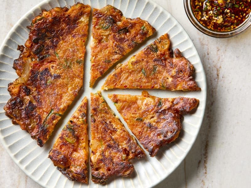 overhead shot of a partially cut kimchi pancake on a white plate, with a bowl of dipping sauce to the side