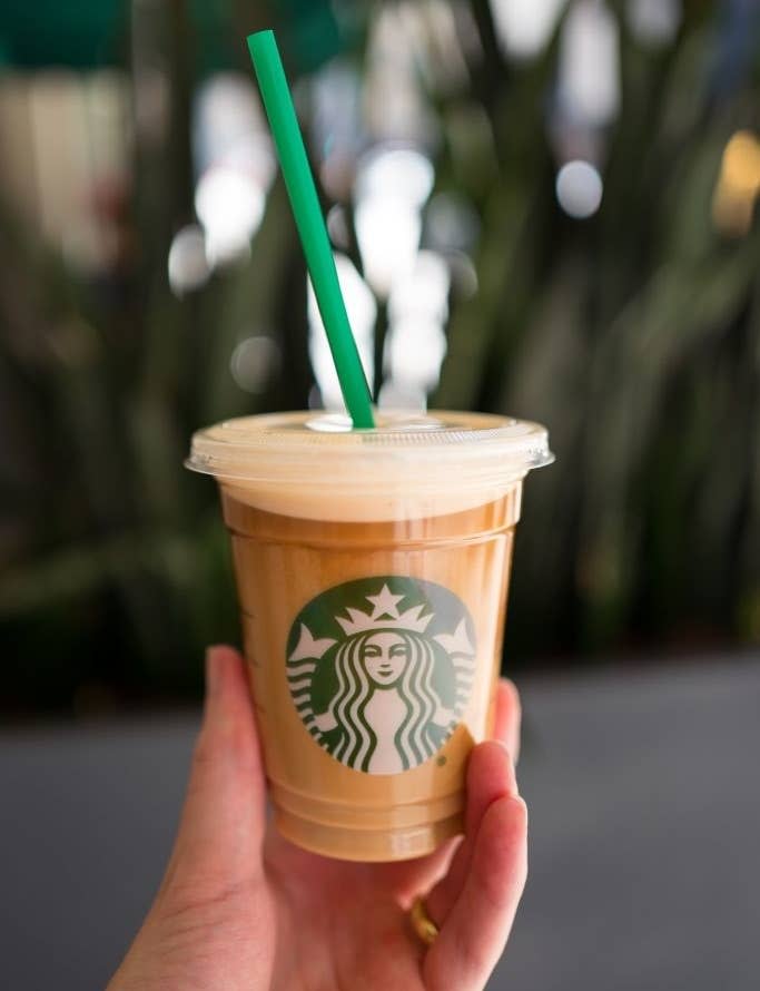 A hand holding a Starbucks iced coffee with a green straw. The Starbucks logo is visible on the cup