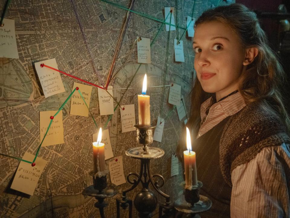 millie bobby brown as enola holmes, standing in front of a map with post-it notes and strings connecting them, illuminating the picture with a candelabra