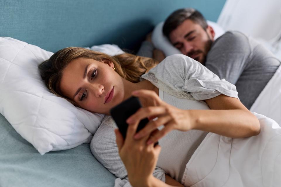 young woman using mobile phone and checking messages in bed while her husband is asleep