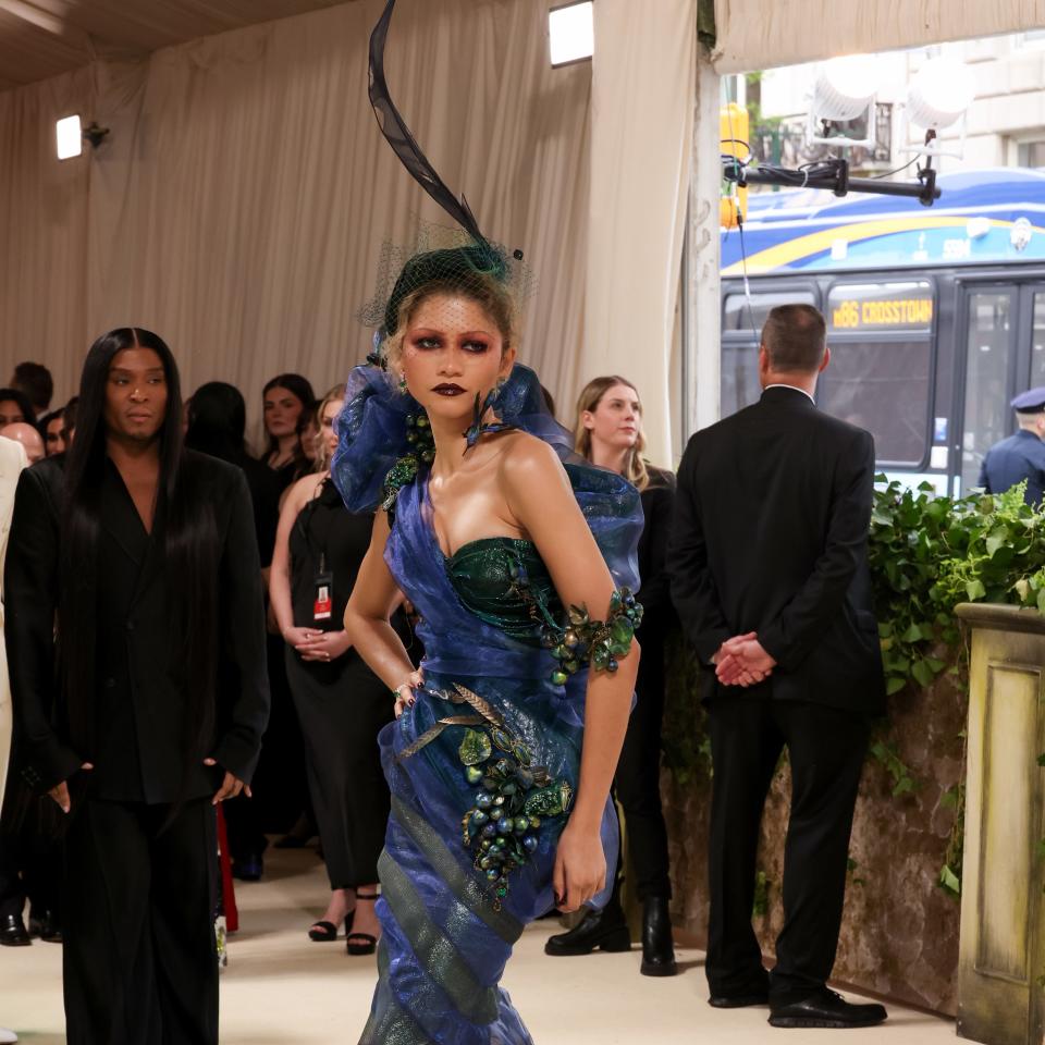 Zendaya in an elaborate blue gown with ruffled details and a dramatic headpiece on a themed event carpet