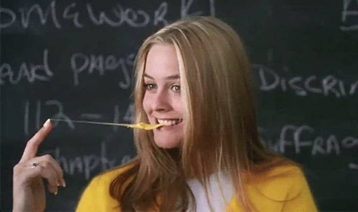 Alicia Silverstone holding a stretched out piece of gum with her finger and smiling, with writing on a blackboard in the background