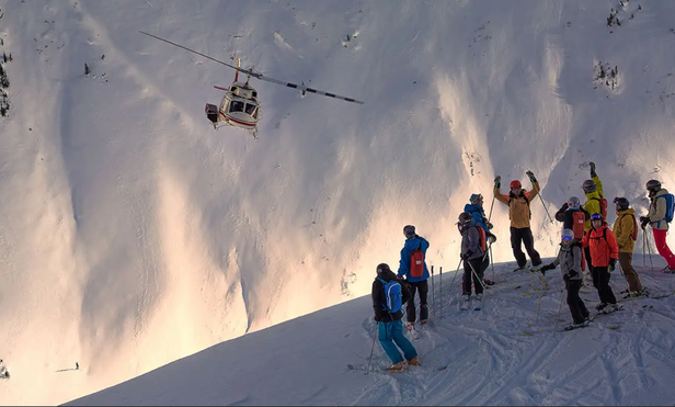 Group of skiiers stand next to helicopter on side of a cliff