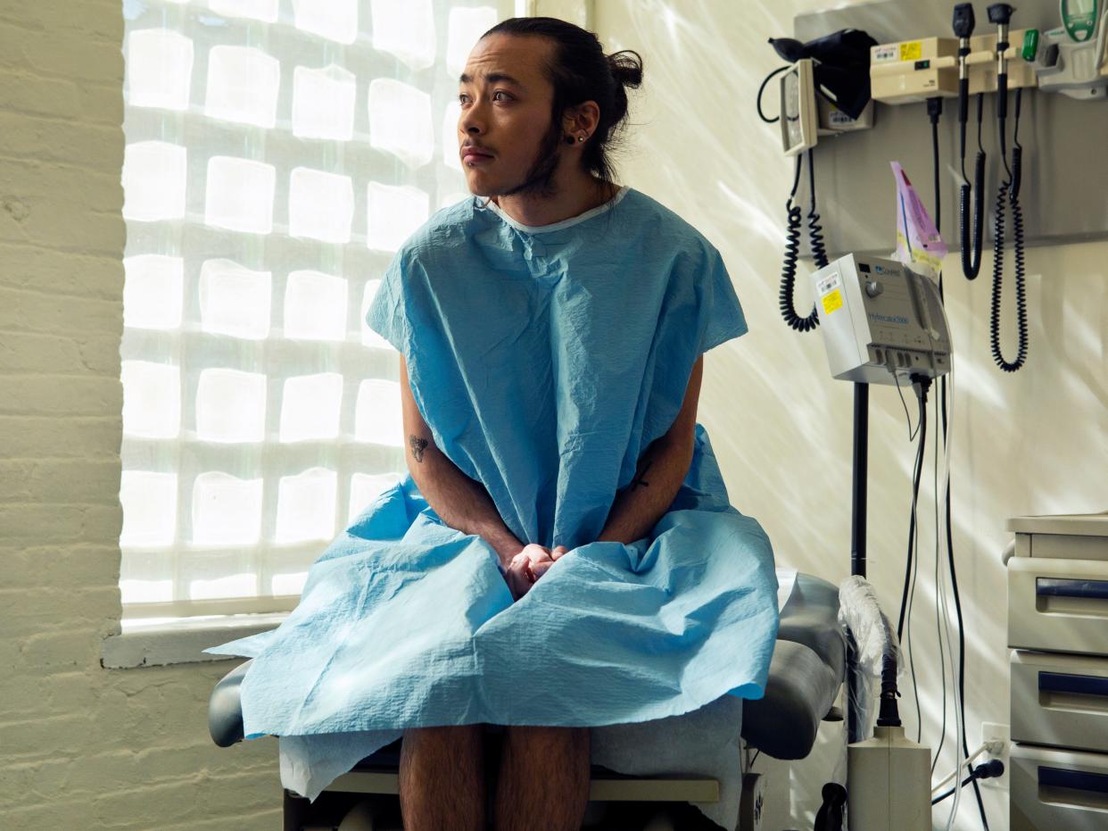 A genderqueer person sitting in a hospital gown sitting in an exam room