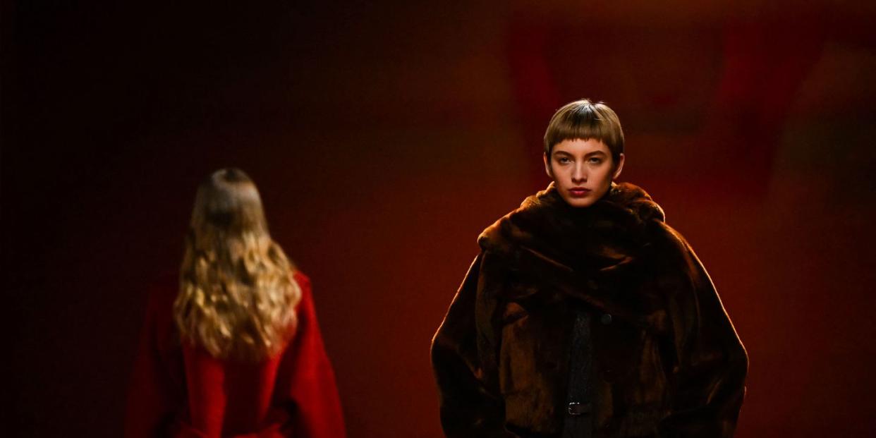 a model with short brown bangs walks towards the camera in a brown coat, while a blond model in a red coat walks in the other direction