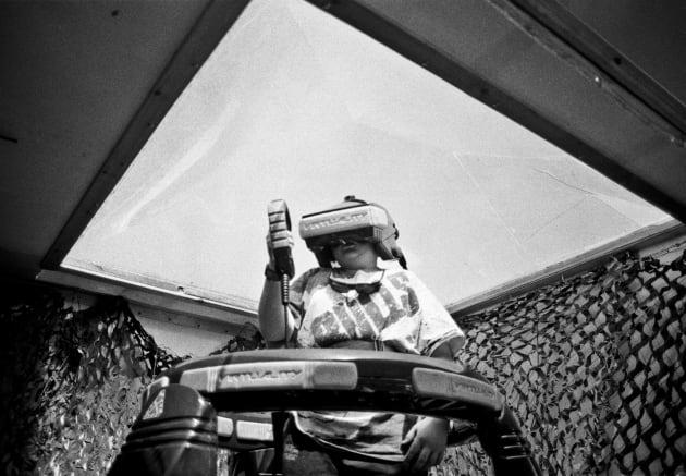 A young boy stands wearing virtual reality mask and hand piece under a plastic roof at a music festival