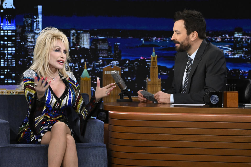 Dolly Parton, in a sparkling dress, talks animatedly with Jimmy Fallon during an interview on his talk show set