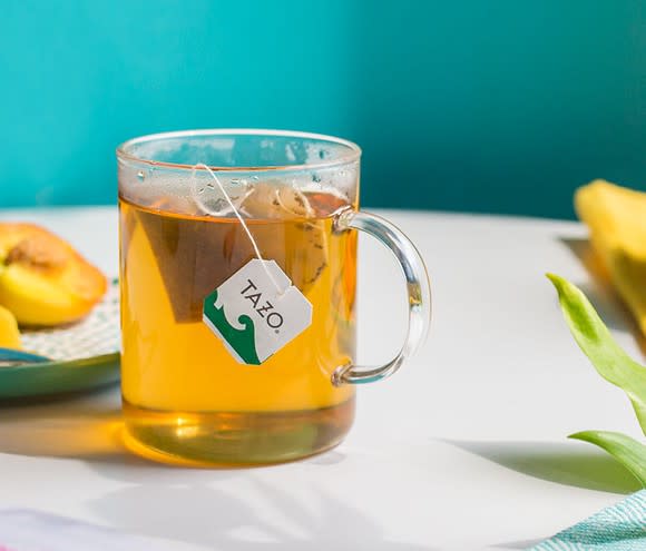 A cup of Tazo tea in a clear glass, sitting on a tablerop near flowers and a plate of fruit.