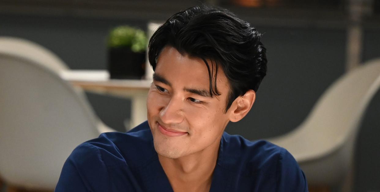 actor alex landi as dr nico kim in season 18 of grey's anatomy, shown in wearing a navy scrub shirt and smiling at someone off camera