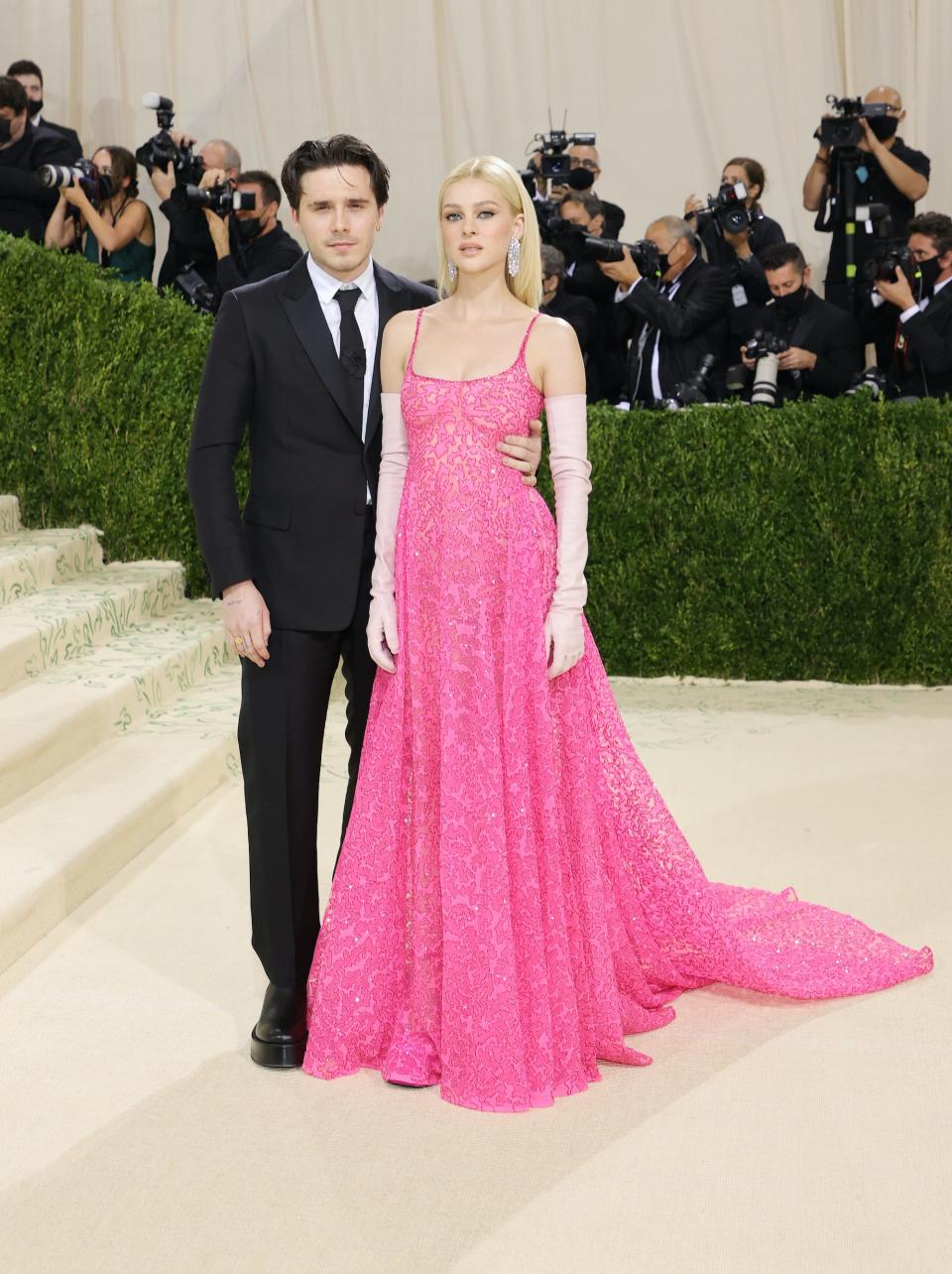 Brooklyn Beckham and Nicola Peltz stand on a cream carpet.