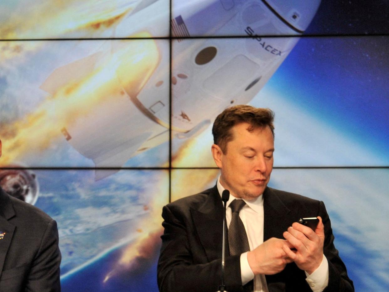 Elon Musk looks at his phone with a display of a space capsule entering orbit behind him.