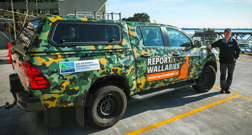 A vehicle painted with camouflage and an anti-wallaby sign. And a council worker standing next to the vehcile.