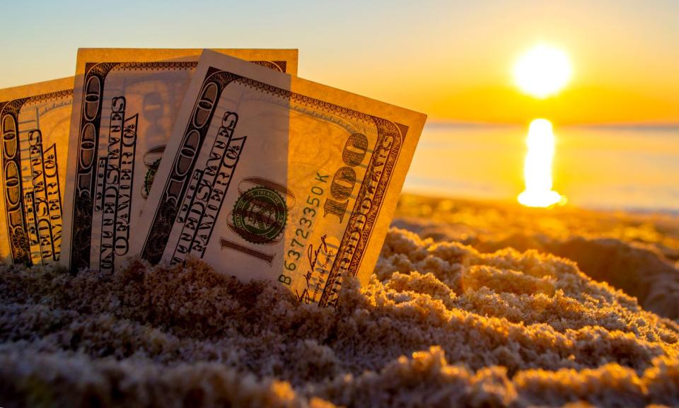 Three fanned one hundred dollar bills partially buried upright in the sand, with the sun rising in the background.