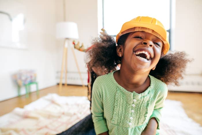 A young child with curly hair, wearing a construction helmet, laughs joyfully inside a bright room with a blanket and a floor lamp in the background