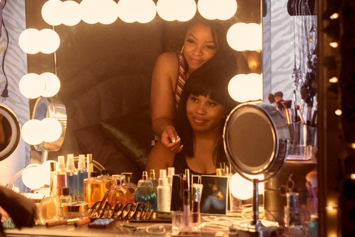 An image of Marissa (Chloe Bailey) smiling while standing behind Dre (Dominique Fishback) in front of a makeup mirror surrounded by lights and cosmetics.