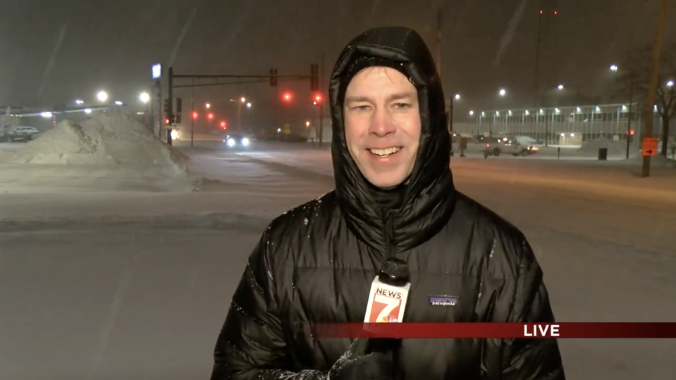 Woodley stands in a black winter coat holding a KWWL microphone outside in the dark with snow falling. (Courtesy Mark Woodley / KWWL)