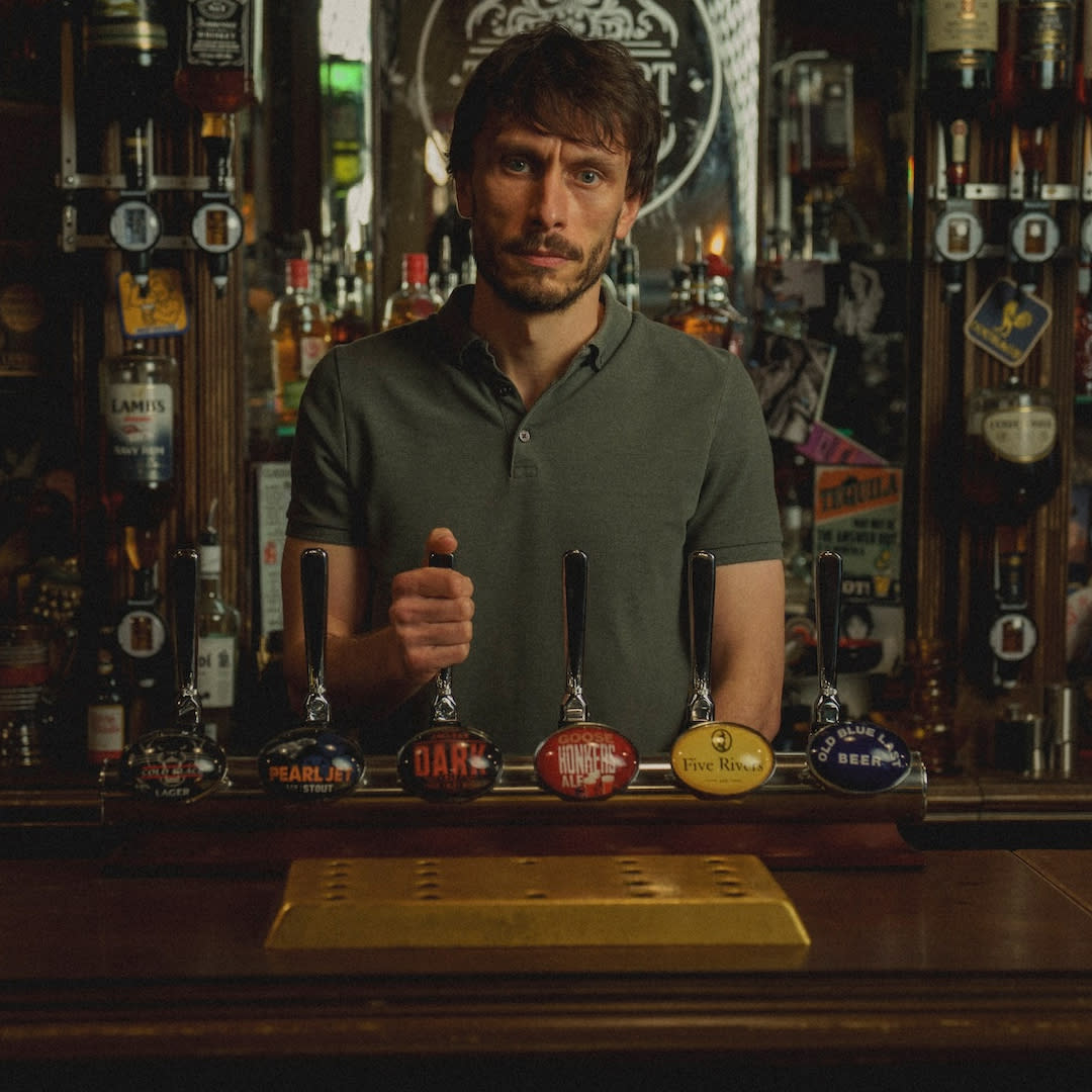  A man (richard Gadd as donny dunn) stands behind a bar with his hand on a tap handle, in netflix's 'baby reindeer'. 