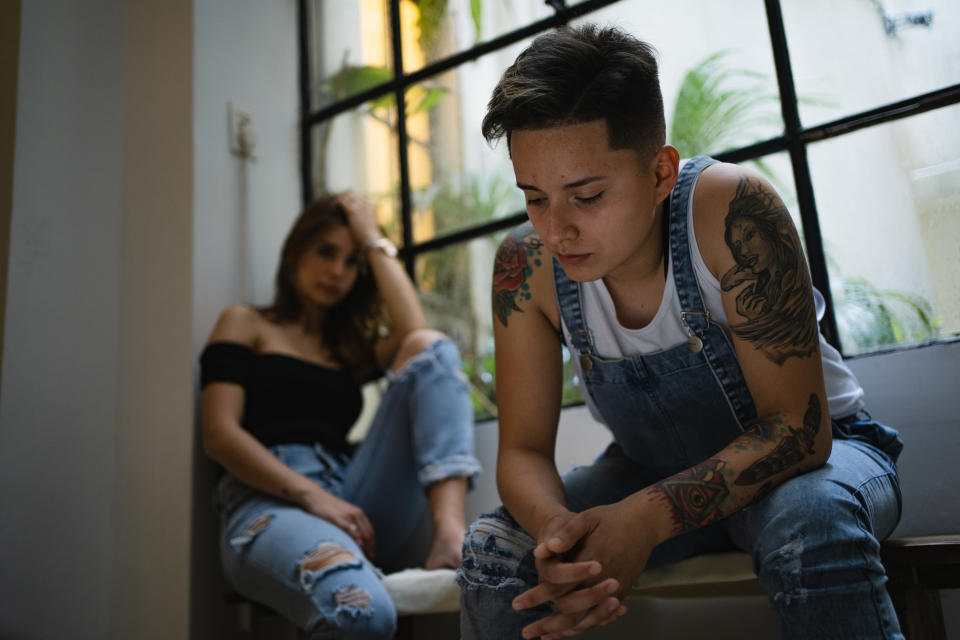 Two people sit by a window. The person in the foreground, with tattoos and short hair, wears a white tank top and blue overalls. Another person sits behind them