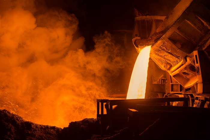 A big steel drum pours hot molten steel into another and steam arises around it in a dark plant.