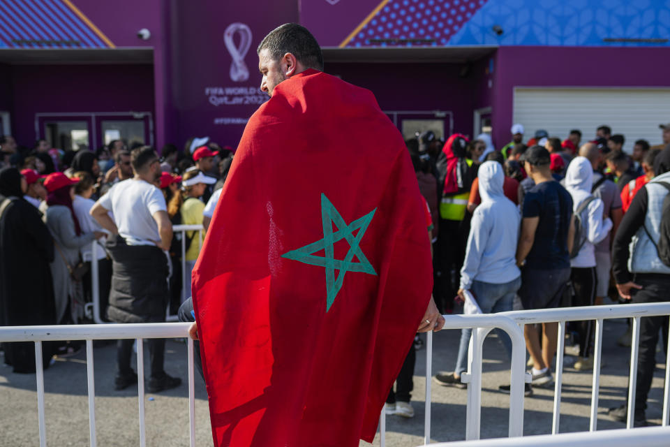 A man draped in a Moroccan flag queues with supporters outside the Al Janoub Stadium in Wakrah, Qatar on Tuesday, Dec. 13, 2022 in the hope of getting tickets for Wednesday's World Cup semifinal soccer match between France and Morocco. (AP Photo/Francisco Seco)