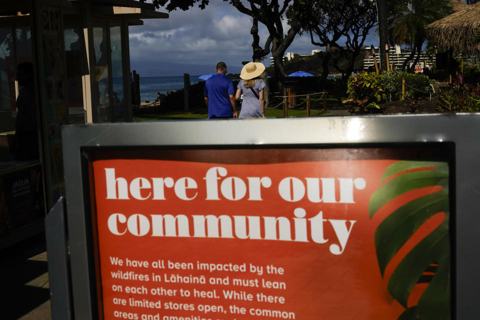 A couple walks by a sign acknowledging the August wildfires at Whalers Village, Wednesday, Dec. 6, 2023, on Kaanapali Beach in Lahaina, Hawaii. Residents and survivors still dealing with the aftermath of the August wildfires in Lahaina have mixed feelings as tourists begin to return to the west side of Maui. (AP Photo/Lindsey Wasson)