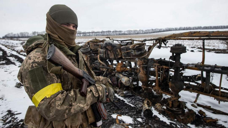 A Ukrainian serviceman with a rifle and yellow armband surveys snowy fields.