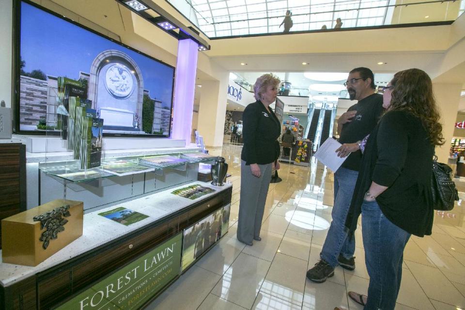 In this photo taken Thursday, Jan. 30, 2014, Forest Lawn regional sales manager Hilda Carabes, left, shows Mark Sanchez and his wife the Forest Lawn stand at the Glendale Galleria mall in Glendale, Calif. Forest Lawn, famous as the final resting place for everyone from Al Jolson to Michael Jackson, has begun staffing outlets at shopping malls, reasoning that planning for death, either for a loved one or yourself, might not be quite as intimidating for some people if it takes place in a lively, happy place like a mall rather than the more somber confines of a cremation home. (AP Photo/Damian Dovarganes)