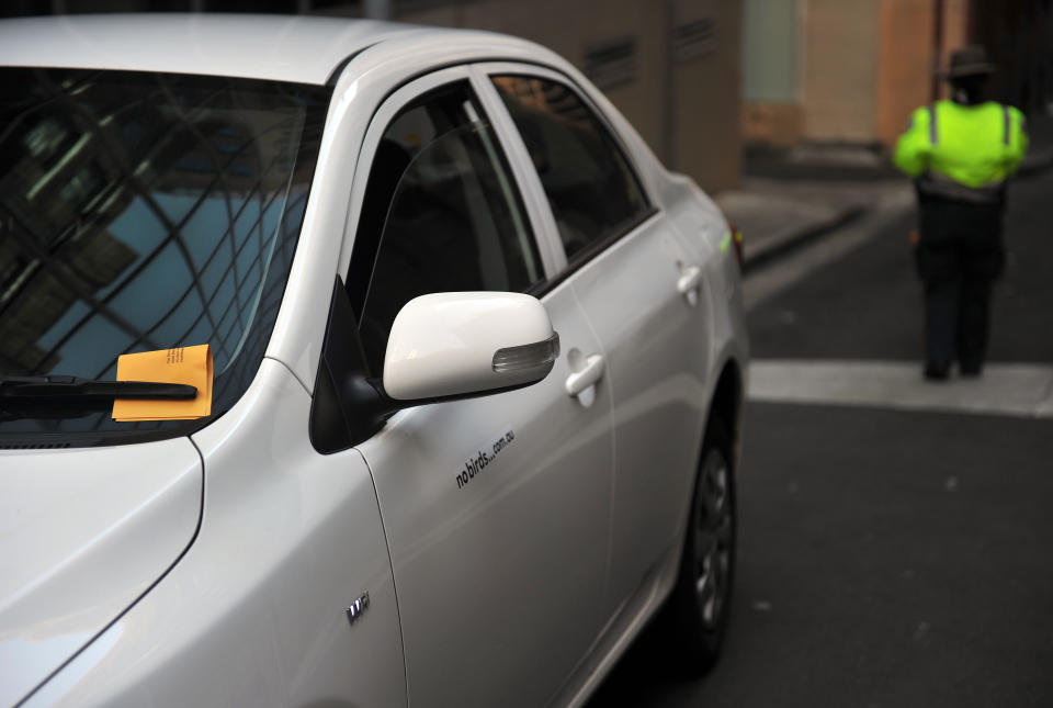 A stock image of a car with a ticket under the windscreen wiper, and a parking officer in the background.