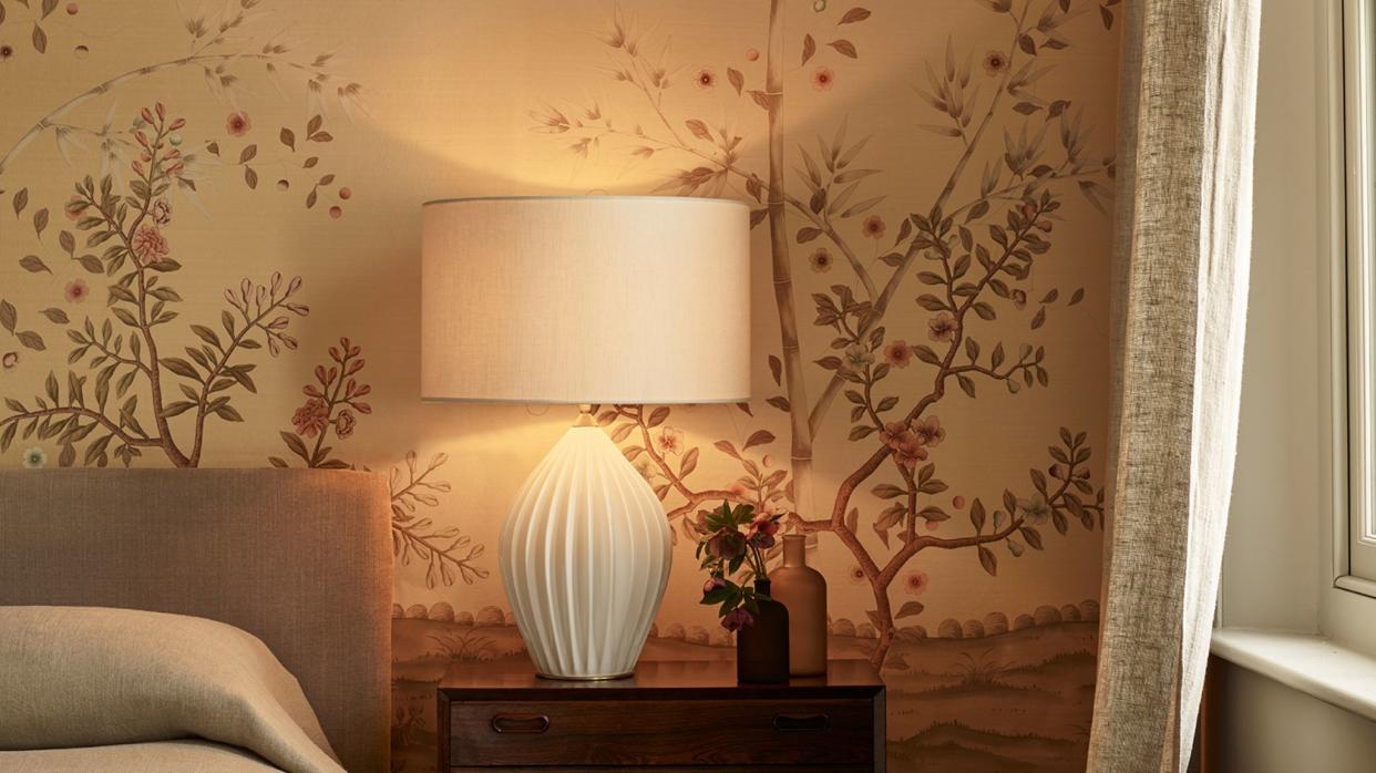  Bedside ceramic lamp with cream base and shade, wooden bedside table and floral wallpaper. 