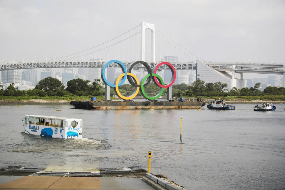 An amphibious bus wades through water for a test cruise as workers on tugboats prepare to move a symbol installed for the Olympic and Paralympic Games Tokyo 2020 on a barge away from its usual spot off the Odaiba Marine Park in Tokyo Thursday, Aug. 6, 2020. The five Olympic rings floating on a barge in Tokyo Bay were removed on Thursday for what is being called “maintenance,” and officials says they will return to greet next year's Games. The Tokyo Olympics have been postponed for a year because of the coronavirus pandemic and are to open on July 23, 2021. The Paralympics follow on Aug. 24. (AP Photo/Hiro Komae)