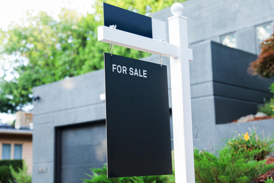 Real estate sign with "For Sale" text in front of a modern house with a garage and landscaped greenery