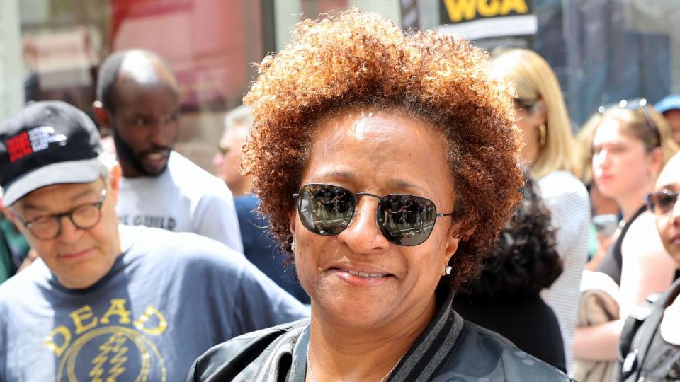wanda sykes smiles at the camera as she stands on a crowded street, she wears a black camo jacket, black shirt and black sunglasses