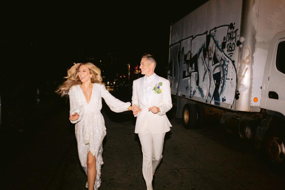 A bride in a white long sleeve dress and a bride in a white suit run down a street holding hands.