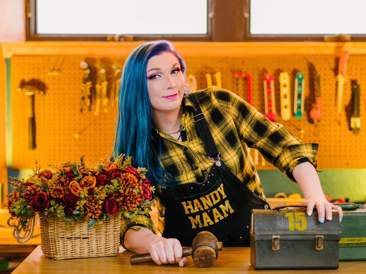 Photo showing Mercury Stardust wearing an apron that says "Handy Ma'am" and leaning on a work bench.