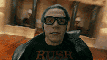 Evan Peters as Quicksilver runs quickly in an indoor scene, wearing goggles, a dark cape, and a Rush t-shirt