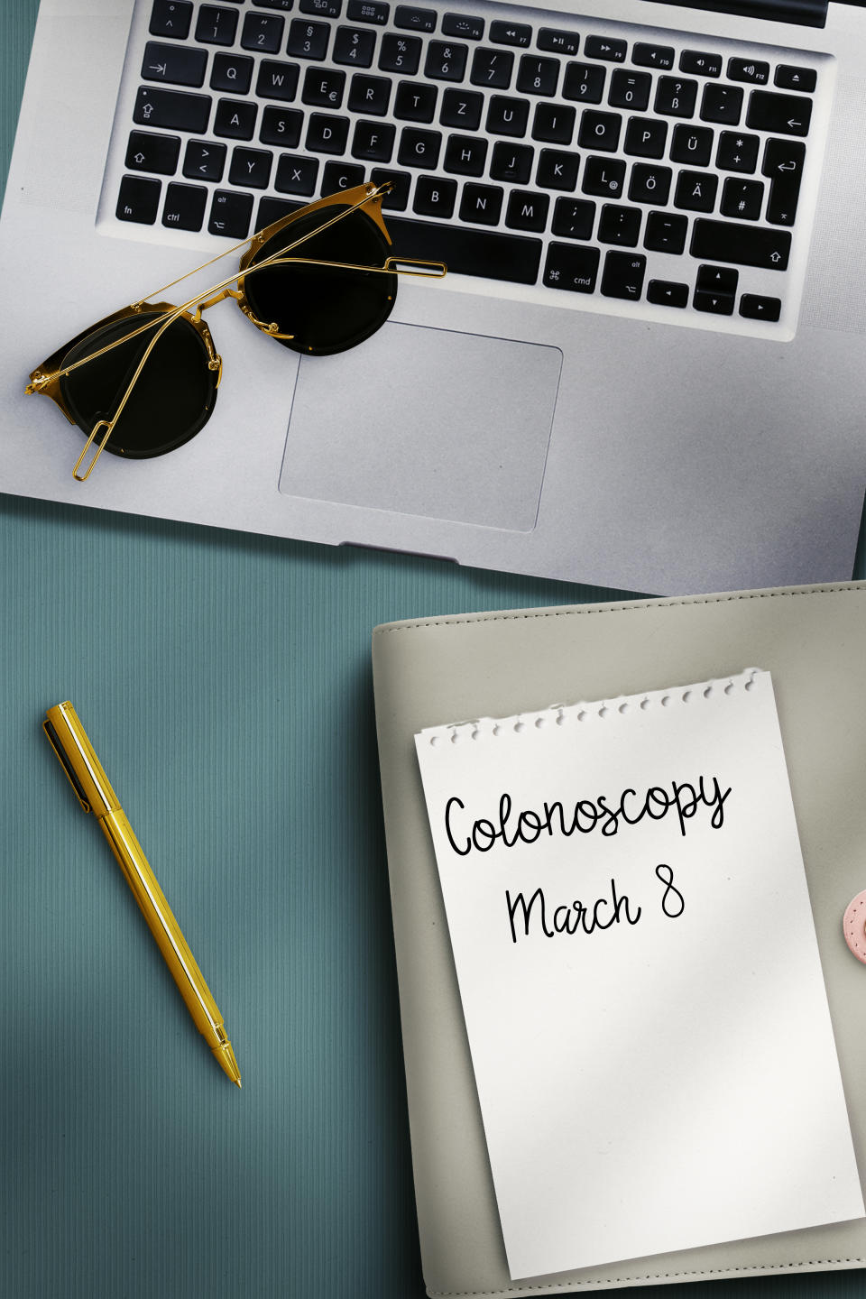 Notepad with handwritten 'Colonoscopy March 8' beside laptop, sunglasses, and pen