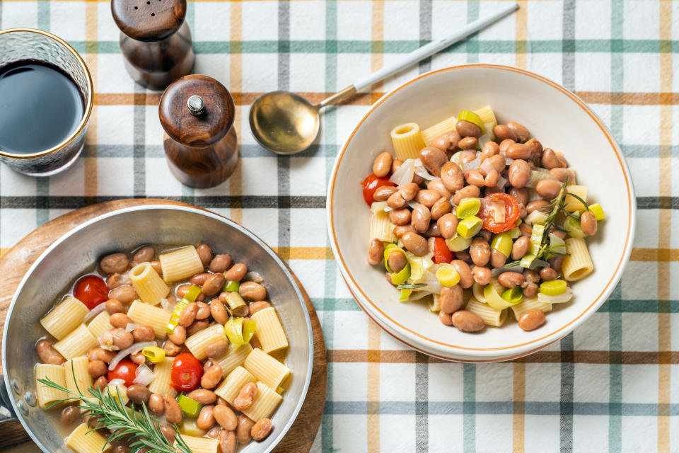 A bowl and pot of bean and pasta soup on a checkered tablecloth, with a spoon and condiments nearby