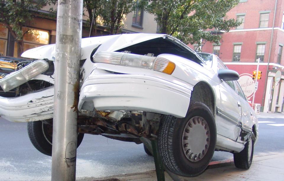 A white car is crashed into a streetlight pole on a city sidewalk with its front lifted off the ground. The impact has caused significant damage