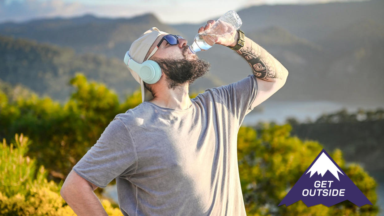  Man drinking from a water bottle while exercising outside surrounded by green trees. 