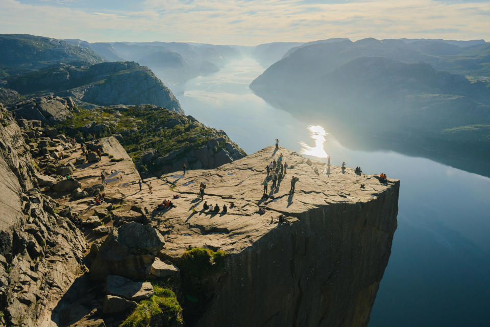 People stand and sit on the large, flat, cliff-top viewpoint of Preikestolen in Norway, overlooking a vast fjord landscape with mountains in the distance