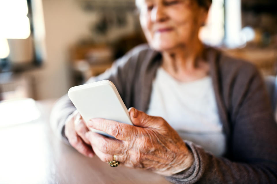 An older adult holding a smartphone, appearing to use it. The background suggests an indoor setting