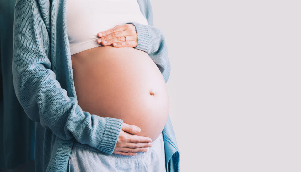 A pregnant person is touching their belly while wearing a sweater and light shirt. No faces are visible