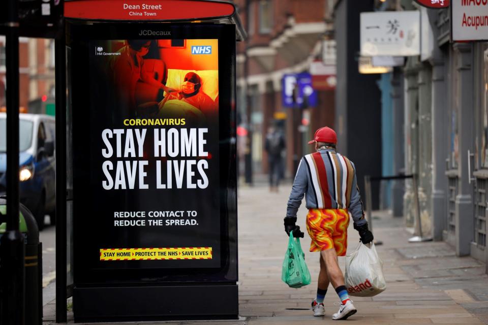 A shopper walks past NHS signage promoting "Stay Home, Save Lives" on a bus shelter in Chinatown, central London