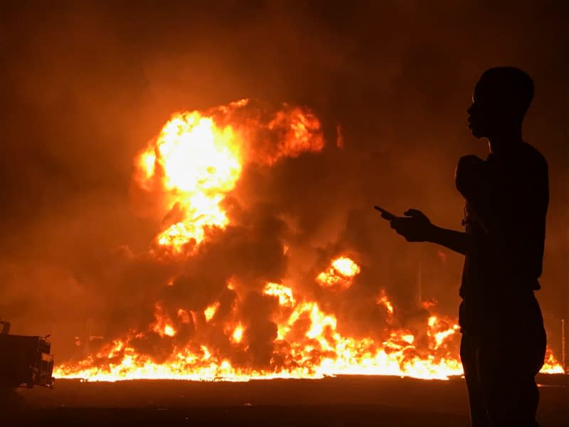 A man stands with his phone in front of flames rising from a pipeline explosion in Abula-Egba, Lagos, Nigeria